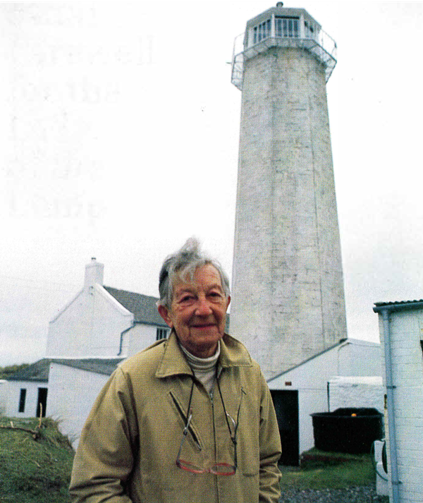 Photo of Peg Braithwaite standing in front of Walney Lighthouse. She has short grey hair and is wearing a beige waterproof jacket over a white polo neck, with a pair of glasses hanging round her neck. The lighthouse is a tall white-painted stone tower in the background.